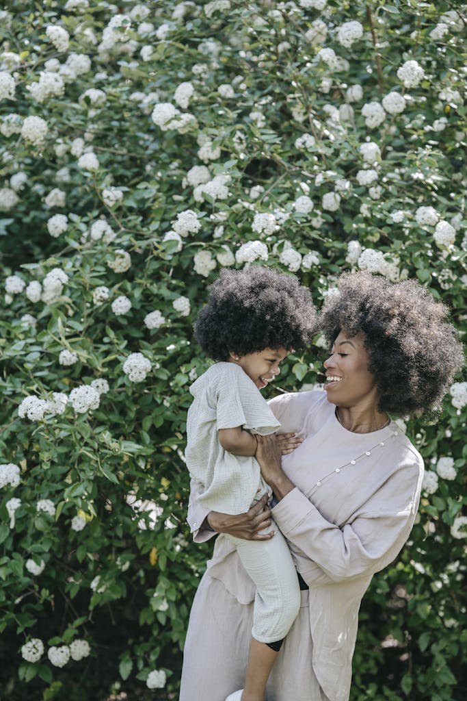 A mother and daughter share a joyful moment surrounded by lush white flowers in a sunny garden.